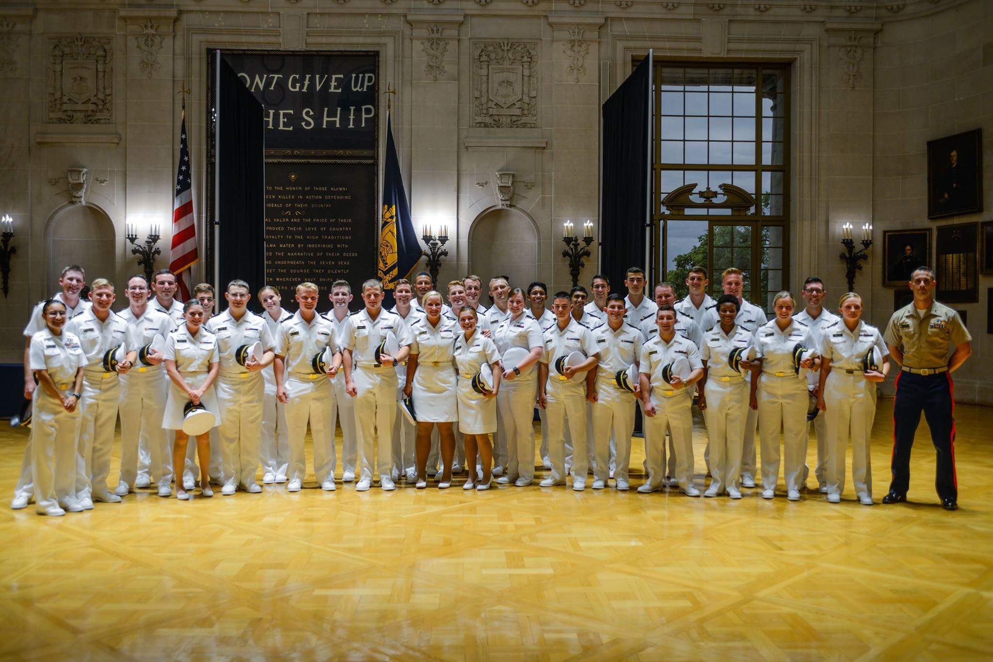 A group of uniformed naval officers and one marine pose for a photo in a large, ornate hall with flags and a “Don’t Give Up the Ship” banner.