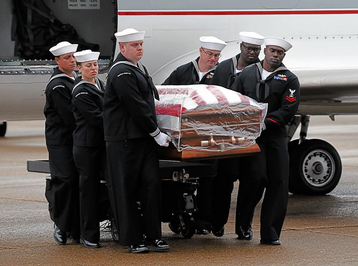 Six Navy sailors carry a flag-draped casket from an aircraft, participating in a formal military ceremony on a wet tarmac.